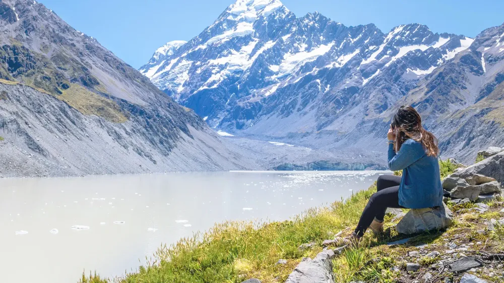 Woman Sits on the Edge of Hooker Glacier Lake with Aoraki Mt. Cook in the Background